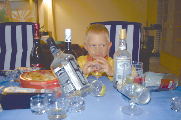 Kind zwischen leeren Gläsern und Flaschen trinkt AlLkohol